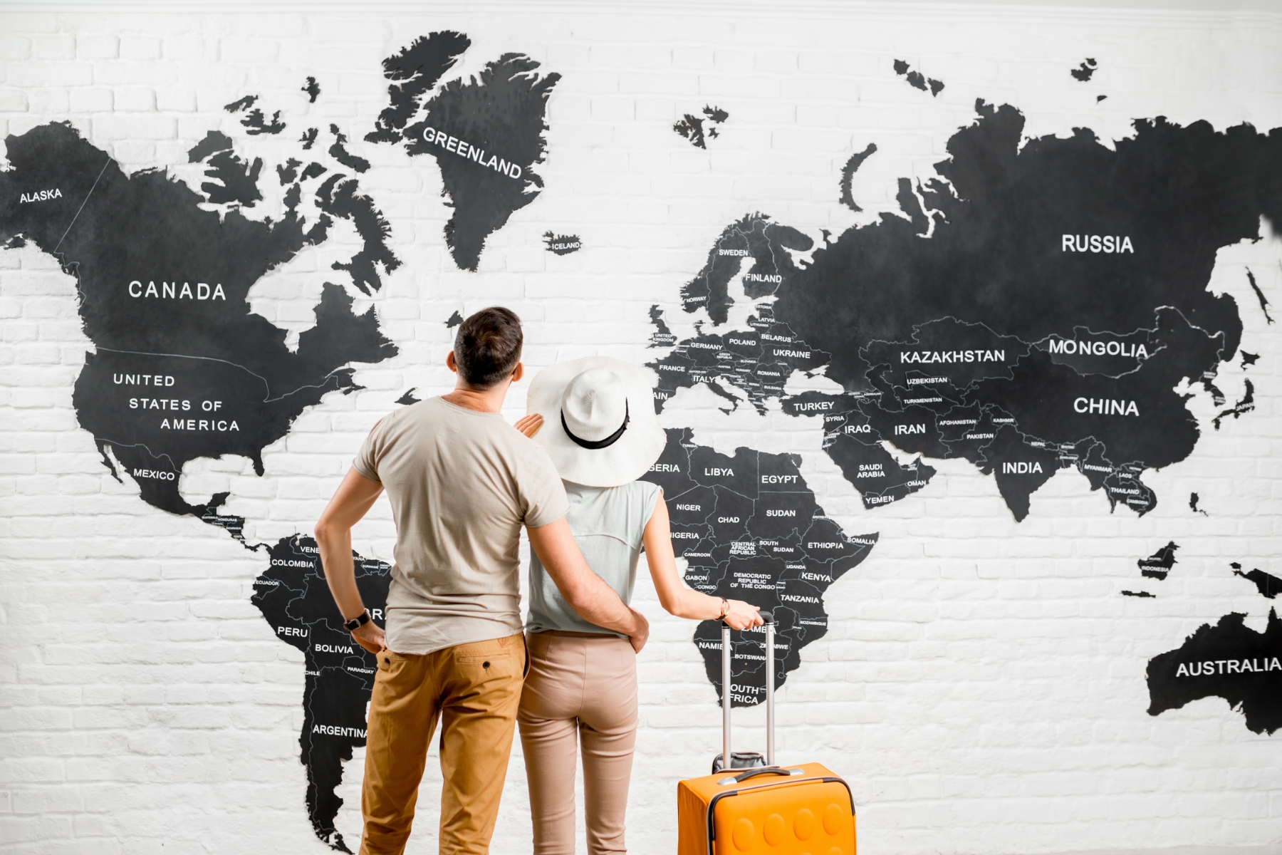 Young couple of travelers standing back near the big world map on the background choosing a place for summer vacation. Illustrative image for FirstMed's Travel Clinic - pre-travel consultancy service.